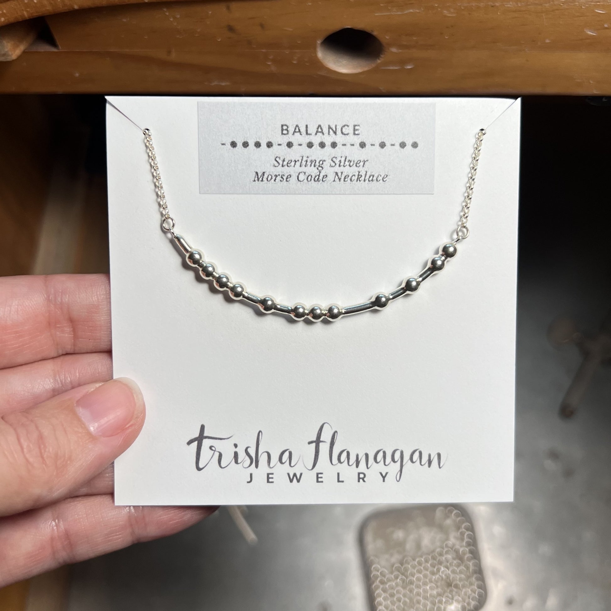Silver Morse Code necklace on a Trisha Flanagan Jewelry card held by a hand.