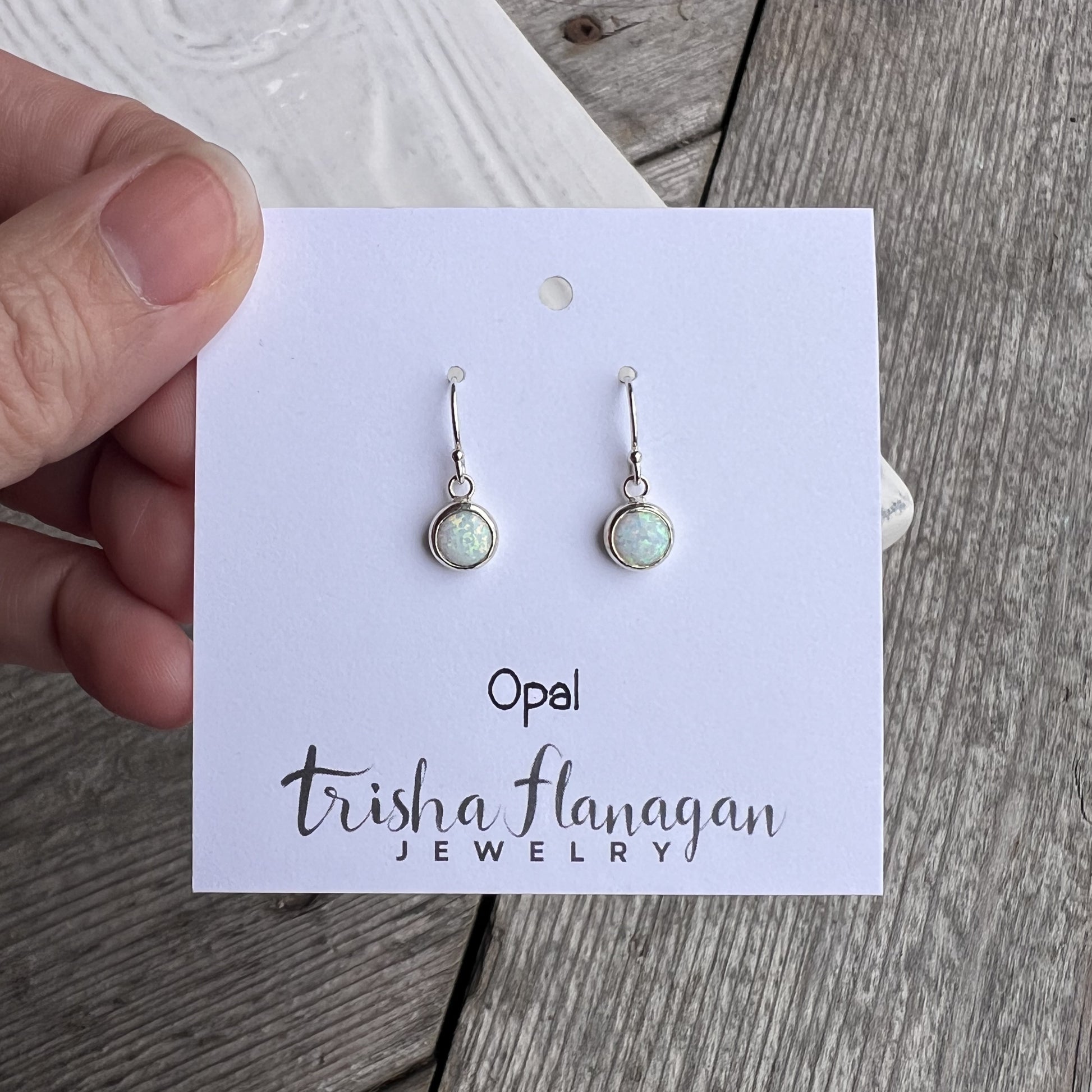 Opal earrings on a Trisha Flanagan Jewelry card held by a hand on a wooden surface.